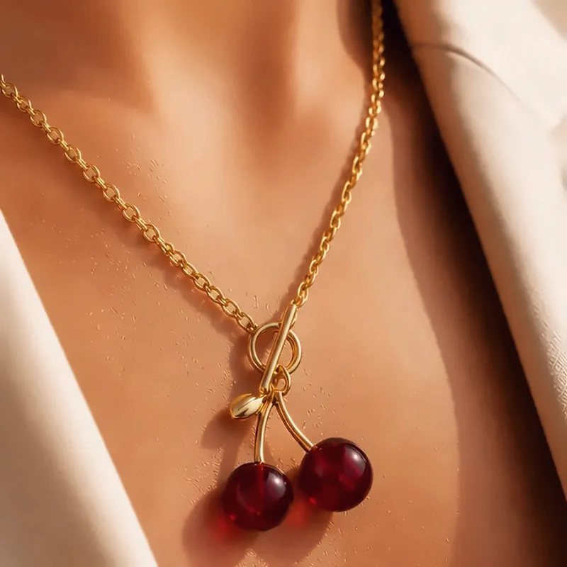 Goldene Halskette mit zwei roten Kirschen als Anhänger, getragen zu einem hellen Blazer.