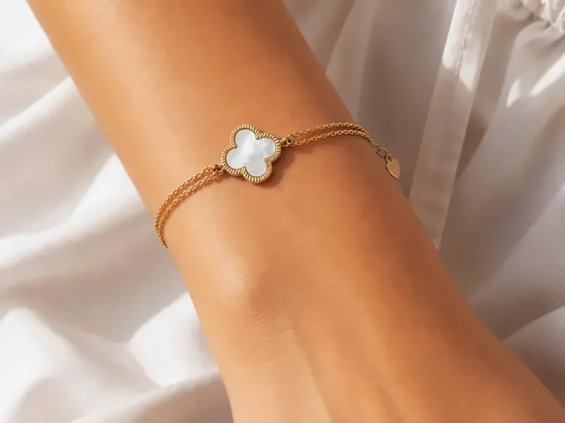 Goldenes Armband mit Kleeblatt aus Perlmutt am Handgelenk, auf weißem Leinen.