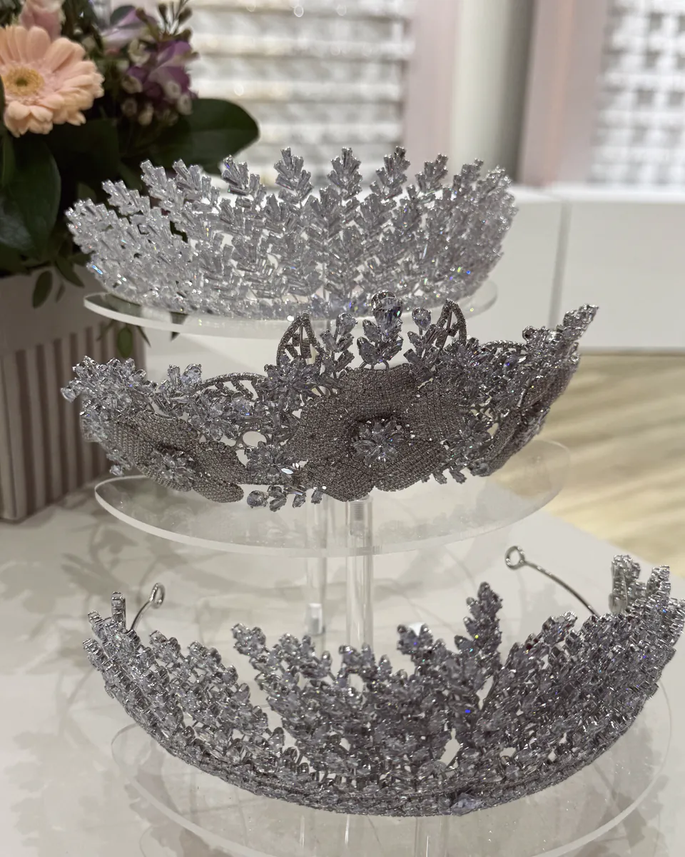 Drei funkelnde Brautdiademe auf einem gläsernen Etagenständer, daneben rosa Blumen.