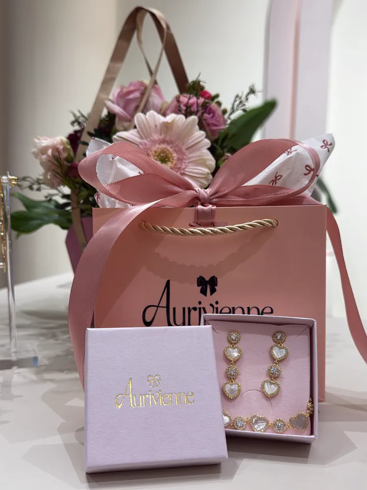 Rosa Aurivienne-Tüte mit Satinschleife und Blumen, davor eine Schmuckbox mit goldenem Logo und Ohrringen mit Herzen.